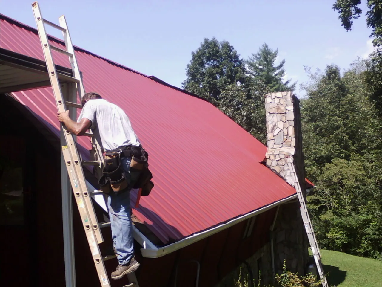 Roofer on a ladder beside a red metal roof and stone chimney — Soffit & Fascia Repair by Roofing Brothers in Sandston